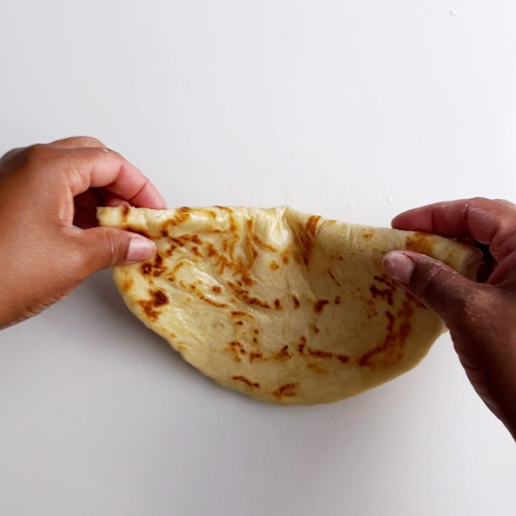 Cooked roti folded in half, held on both ends with finger tips, over a white background