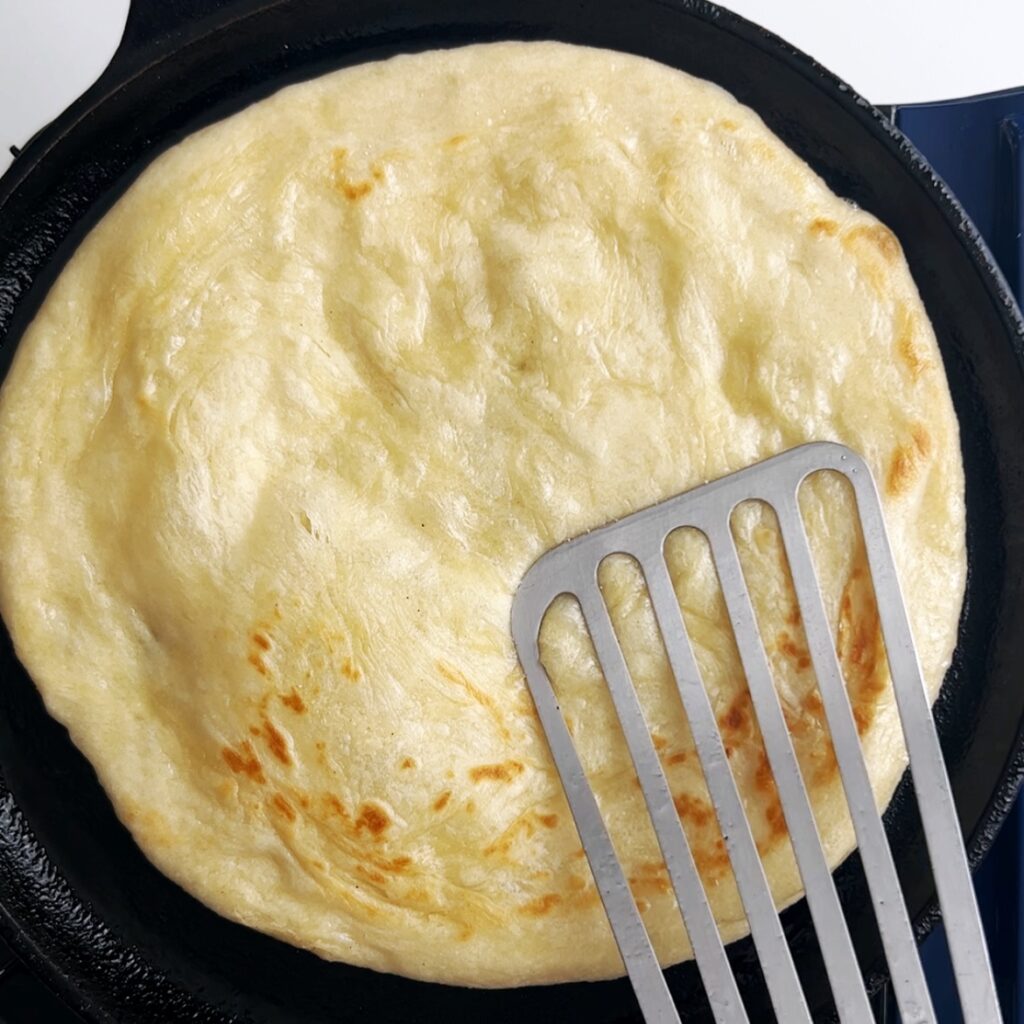 Roti being cooked on a tawa with a spatula pressing down on the roti to create air pockets