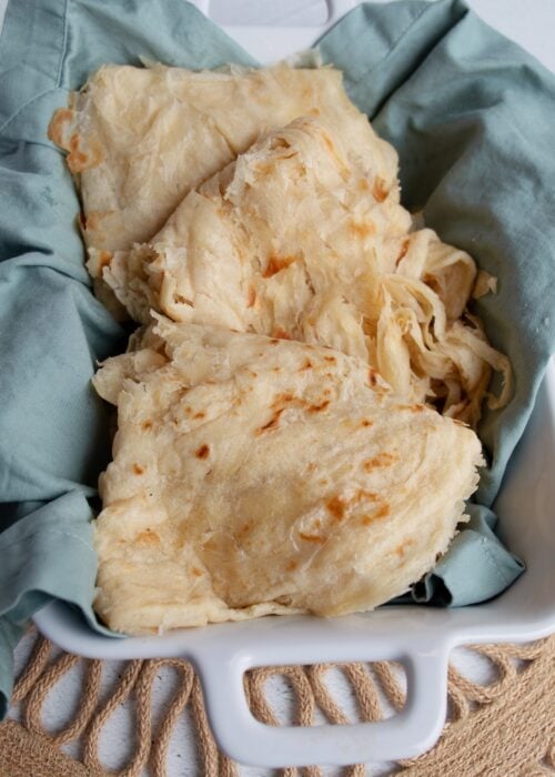 Guyanese roti in a whte rectangular dish. The dish is line with a blueish gray napkin. The dish is resting on a brown, circular macrame place mat set against a white background