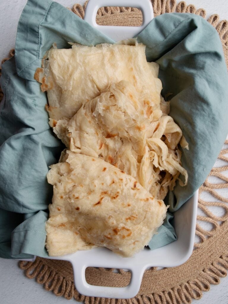 Top down view of Guyanese roti in a white rectangular dish. The dish is line with a blueish gray napkin. The dish is resting on a brown, circular macrame place mat set against a white background