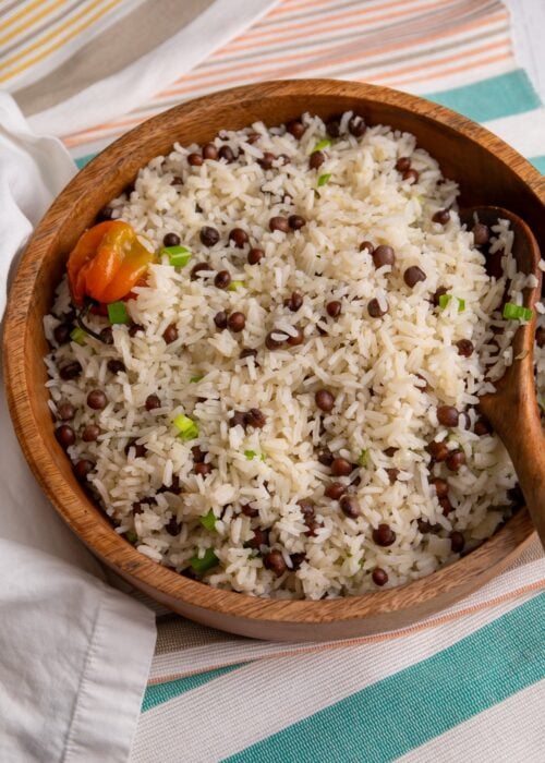 A wooden bowl filled with fluffy Pigeon peas and rice, garnished with chopped scallions and a whole Scotch bonnet pepper on top. The bowl sits on a striped cloth with a wooden spoon resting inside, highlighting the simple, comforting Caribbean rice dish