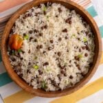 A wooden bowl filled with fluffy Pigeon peas and rice, garnished with chopped scallions and a whole Scotch bonnet pepper on top. The bowl sits on a striped cloth.