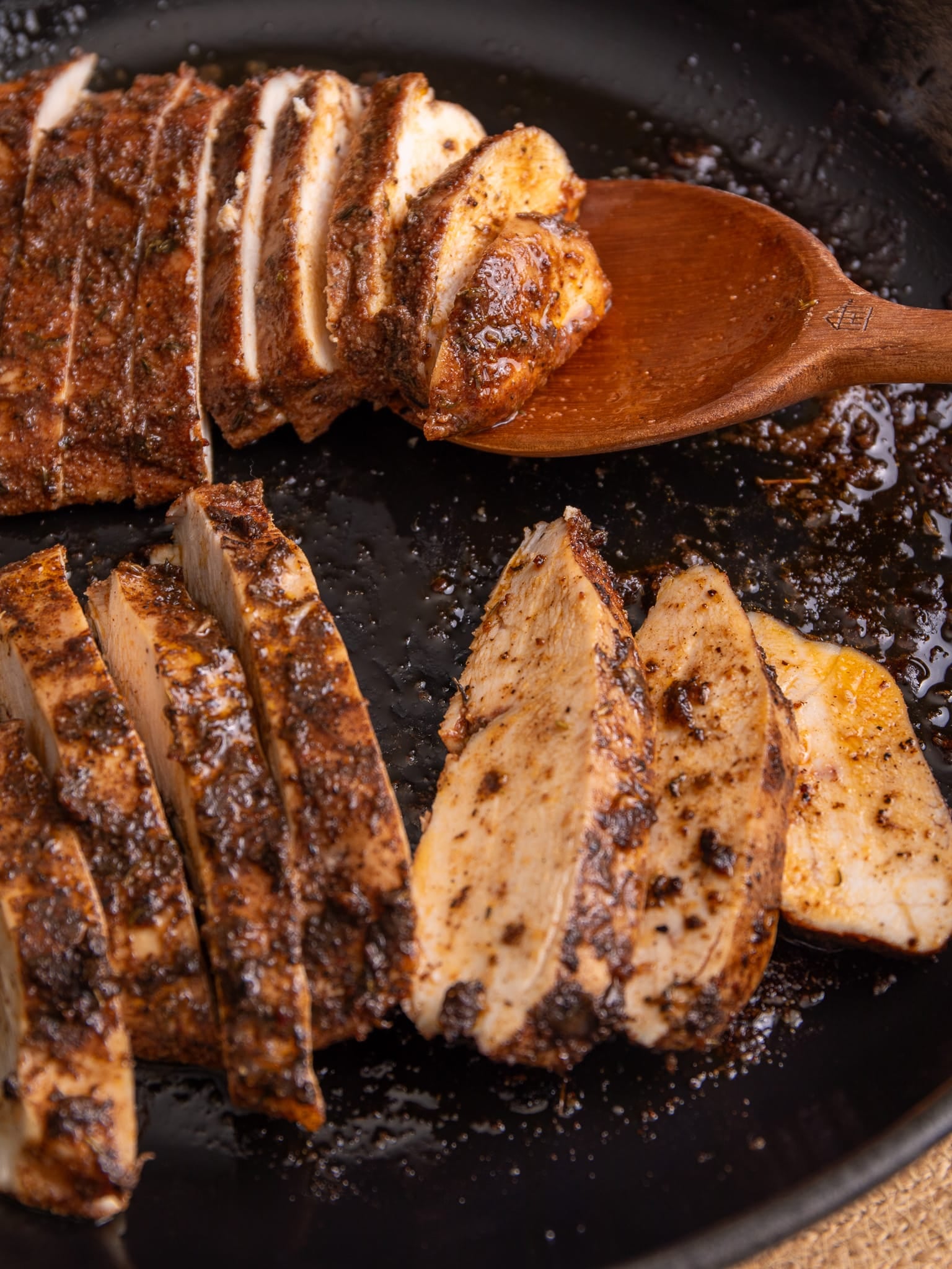 Sliced jerk chicken breast in a cast iron skillet, showing juicy interior and charred seasoning, served on a wooden spoon.