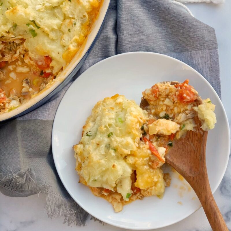 Slice of fish pie on a white plate with a wooden spoon set on a gray napkin