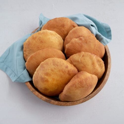 8 golden brown Guyanese bake/s in a wooden circular bowl, lined with a blueish grey cotton napkin.