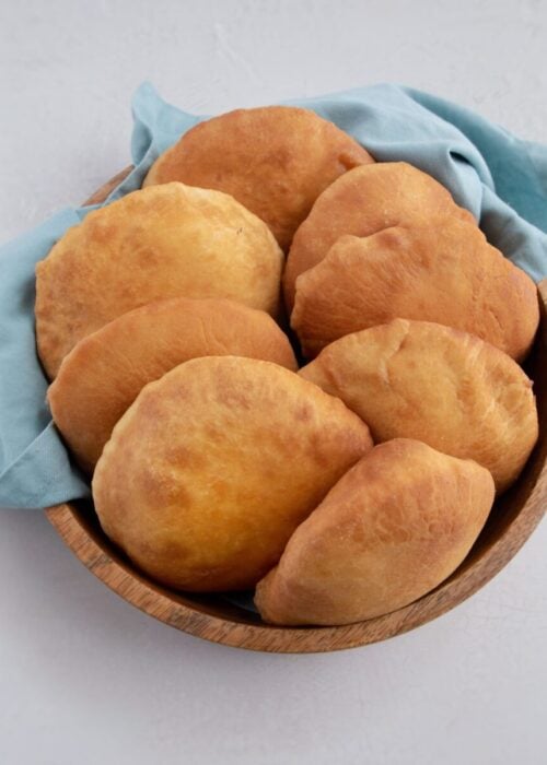 8 golden brown Guyanese bake/s in a wooden circular bowl, lined with a blueish grey cotton napkin.