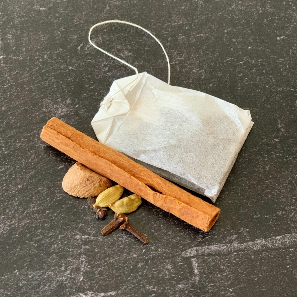 close up view of a black tea bag beside a cinnamon stick, cardamom pods, cloves and nutmeg
