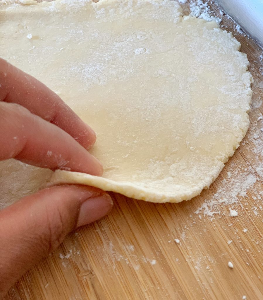 two fingers pinching the side of flattened dough to show how thin it is