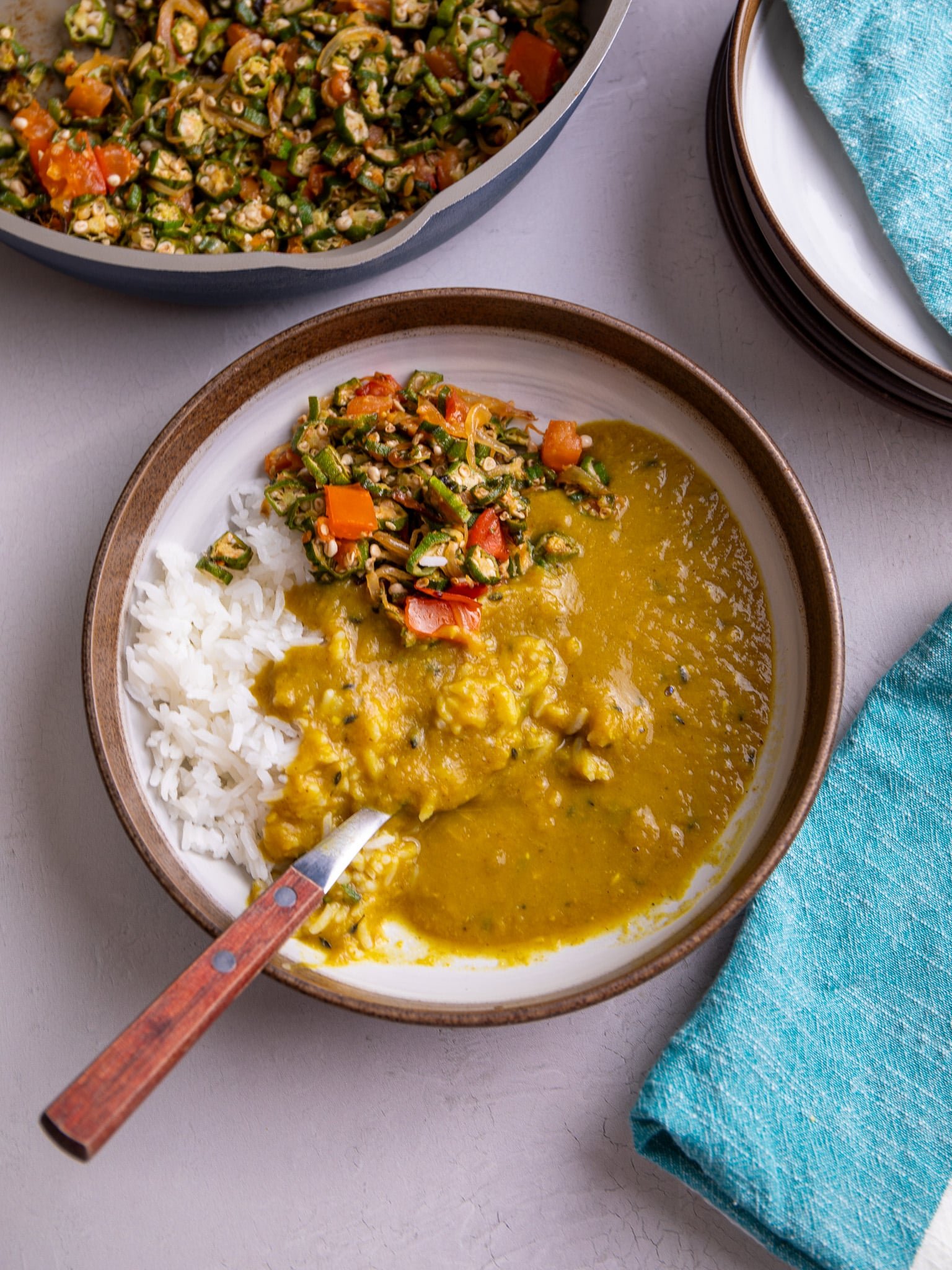 White rice, sautéed okra and dhal is served in a white plate with a brown rim. There is a silver spoon with a wooden handle in the bowl .