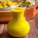 A small glass carafe filled with smooth, bright yellow mango salad dressing speckled with herbs sits on a wooden surface, with a blurred bowl of salad and a wooden tray in the background.