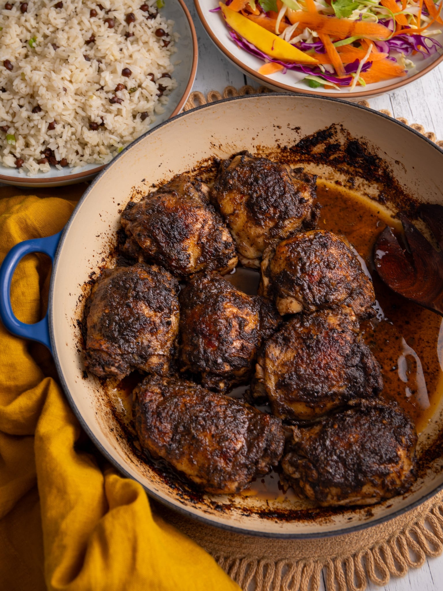Oven-baked jerk chicken thighs in a skillet with pan juices, served with rice and peas and a fresh cabbage and mango slaw on the side.