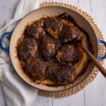 Oven-baked jerk chicken thighs in a skillet with pan juices and a wooden spoon, set on a white wooden table with a linen cloth.