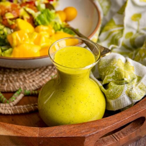 A small glass carafe filled with bright yellow tangy mango vinaigrette sits on a wooden tray, with a fresh salad of lettuce, cherry tomatoes, and mango chunks blurred in the background alongside a green patterned cloth.