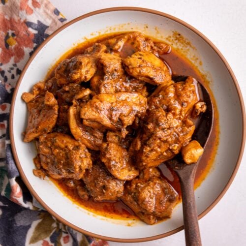 Caribbean Stew Chicken plated in a white bowl with a brown rim. A wooden spoon is scooping up some of the chicken.