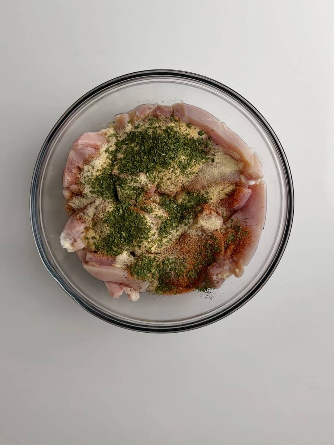 Dried herbs sprinkled over chicken thighs in a glass bowl for Caribbean chicken stew.