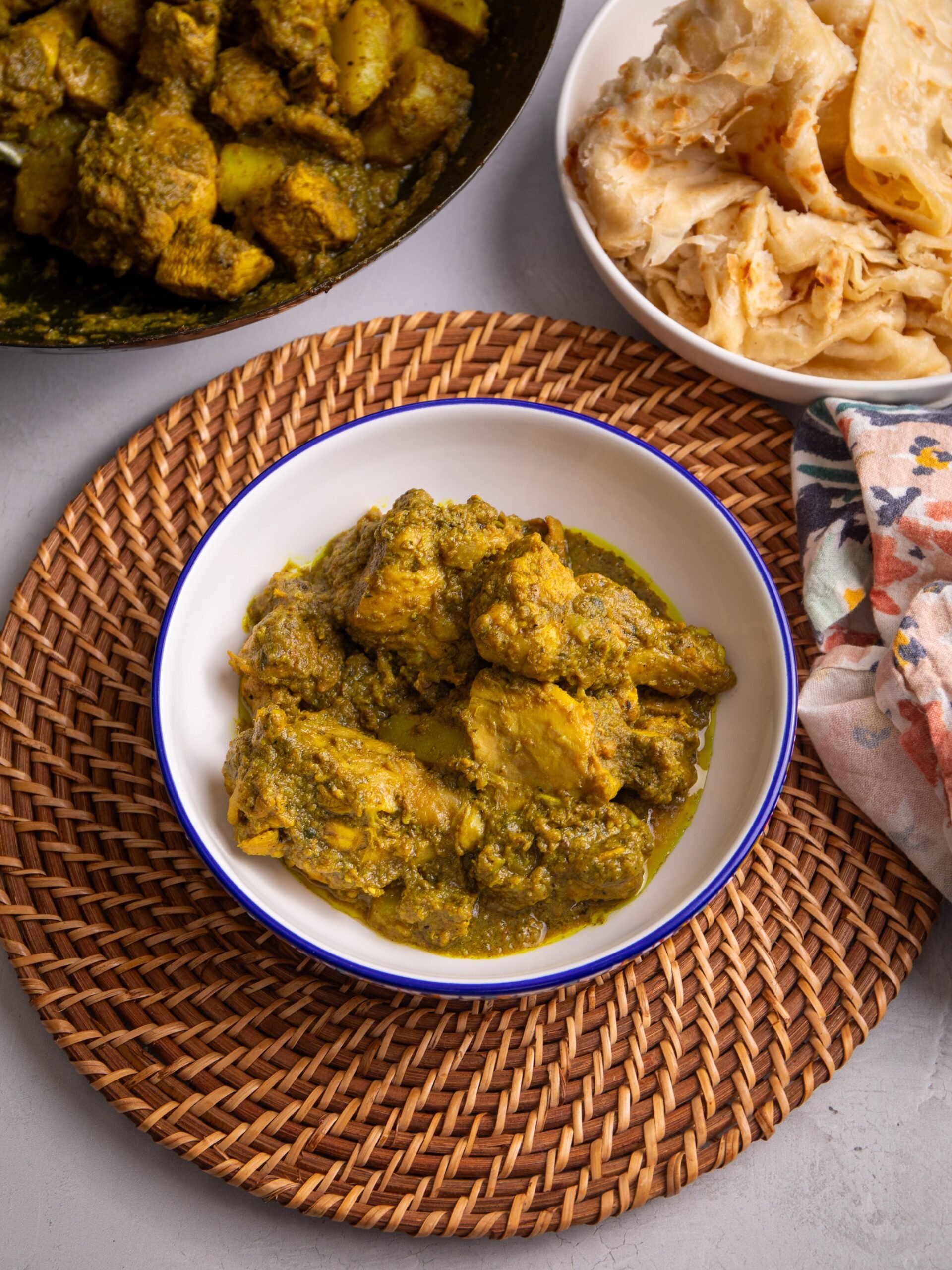 Chicken curry in a white bowl with a blue rim. The bowl is sitting on a woven mat. to the right of the bowl there is a bowl of roti visible and to the top left there is a pot of chicken curry with about 1/3 of the pot visible.