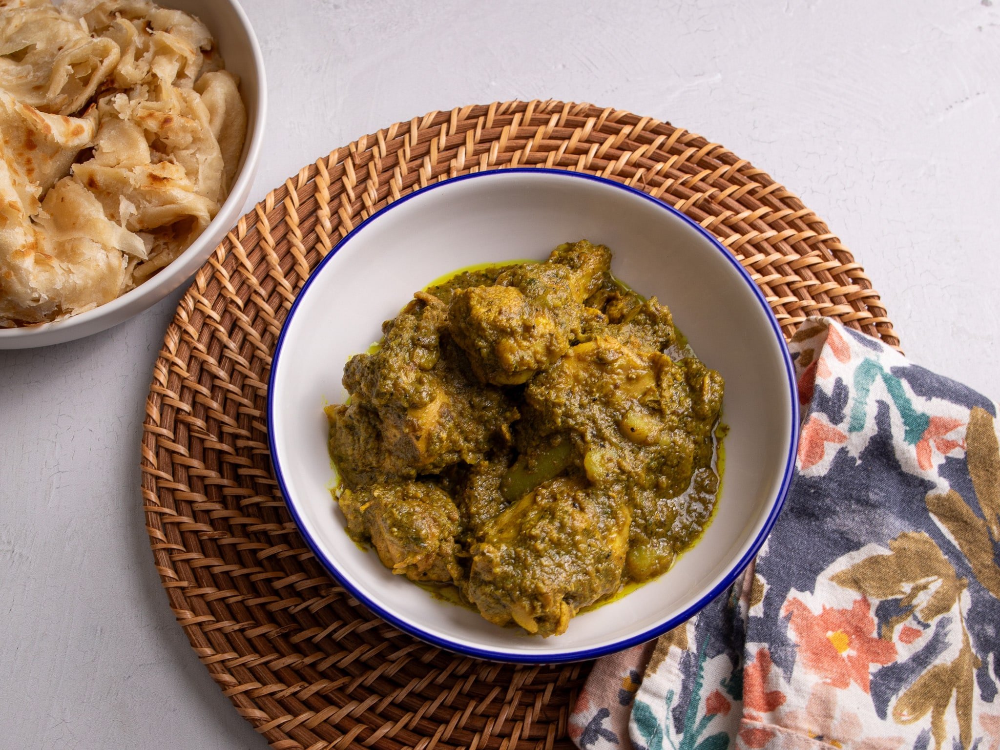 Guyanese Style Chicken Curry in a white bowl with a blue rim on a woven charger plate. A multi-colored napkin is on the bottom right and a bowl with roti is visible on the top left. 
