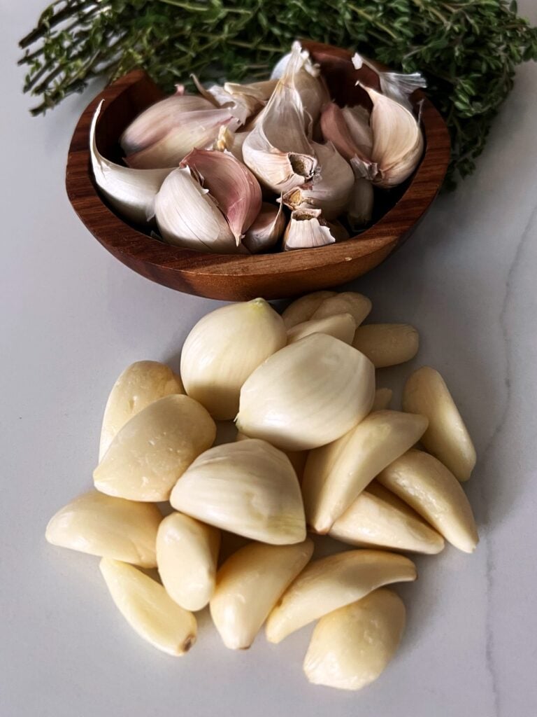 Peeled garlic on a counter with a backdrop of unpeeled garlic in a bowl and thyme stems