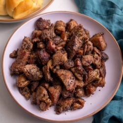 Guyanese garlic pork in a pink bowl with a plate of plait bread to the left of the bowl and a teal napkin to the right