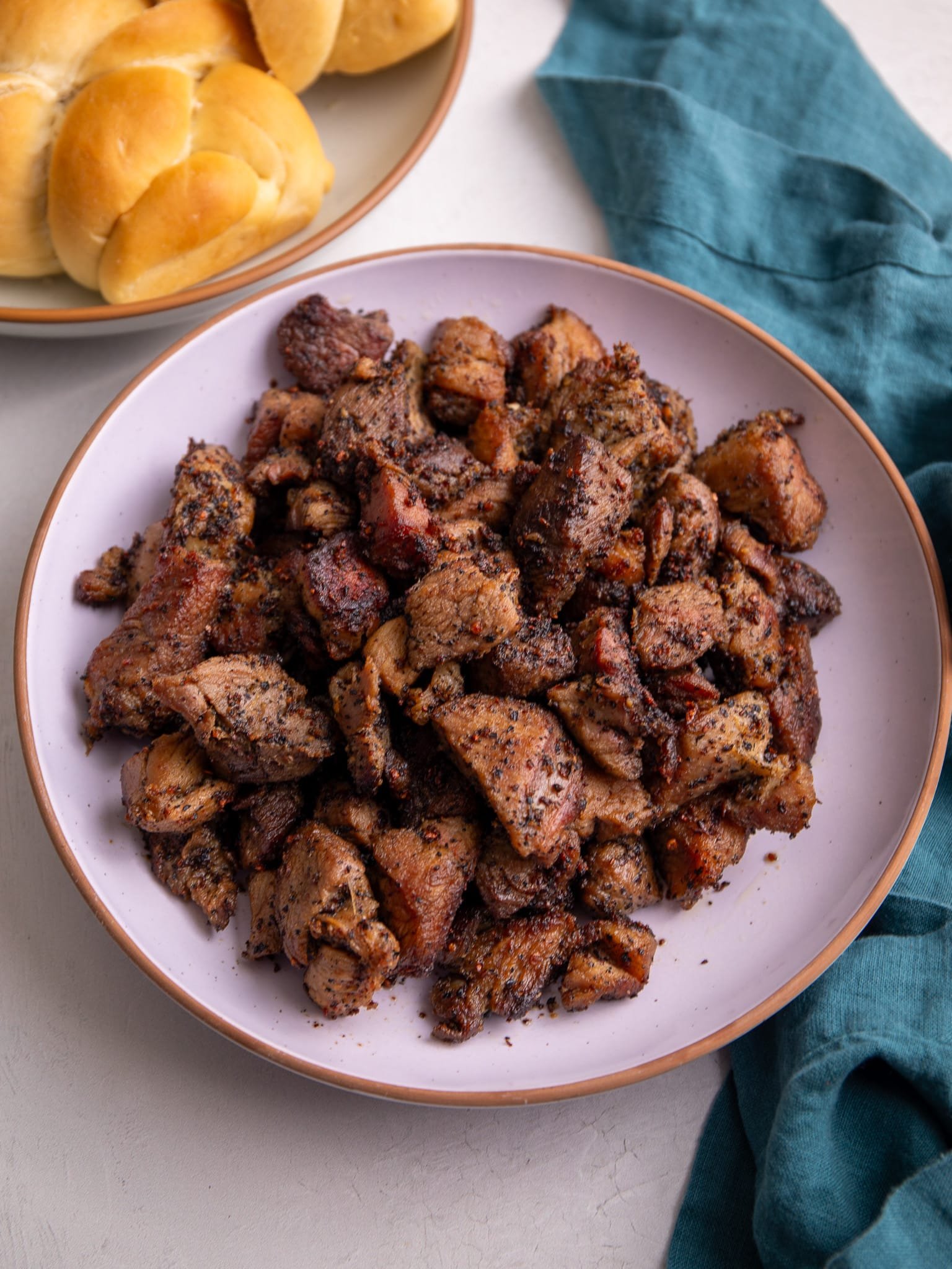 Guyanese garlic pork in a pink bowl with a plate of plait bread to the left of the bowl and a teal napkin to the right