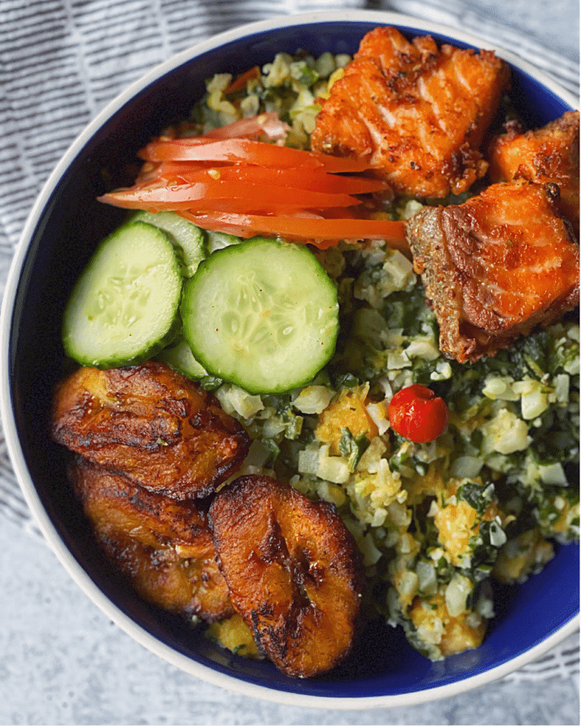 a bowl of cauliflower cook up rice  topped with fried fish, plantains, and fresh veggies