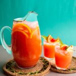 Caribbean rum punch served in a large glass pitcher with tropical garnishes and two glasses over ice