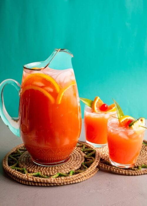 Caribbean rum punch served in a large glass pitcher with tropical garnishes and two glasses over ice