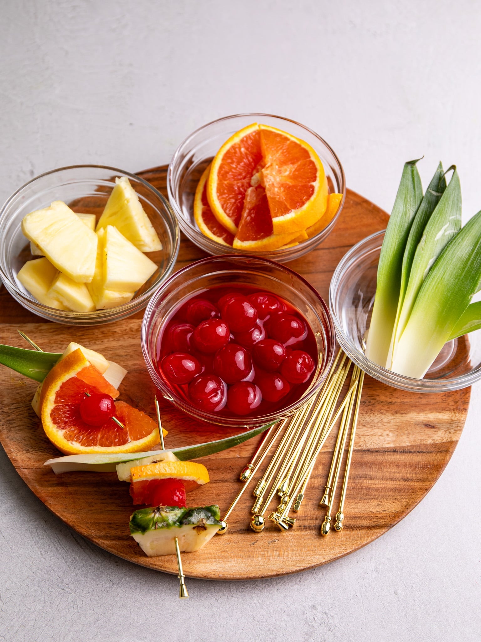Tropical rum punch garnishes including maraschino cherries, orange slices, pineapple wedges, and pineapple leaves arranged on a wooden board with cocktail picks.
