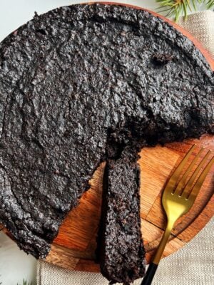 A black cake with a single slice on a wooden plate with a gold and black spoon