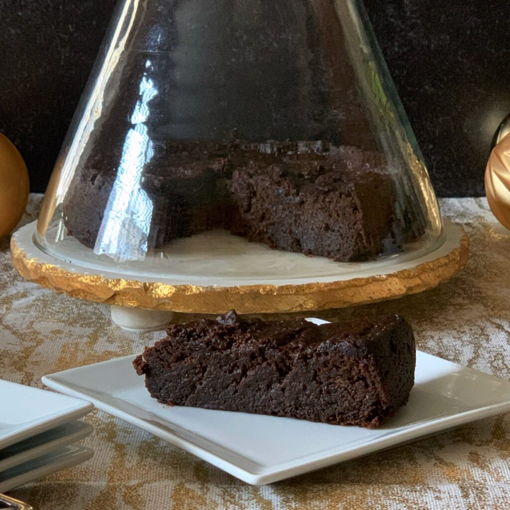 A slice of black cake on a white plate with the remainder of the cake in the background