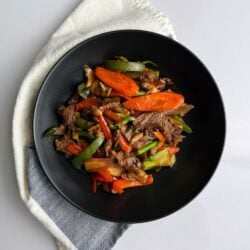 Steak and veggies stir fry in a black bowl on a white and gray napkin