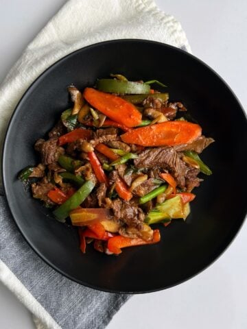 Steak and veggies stir fry in a black bowl on a white and gray napkin