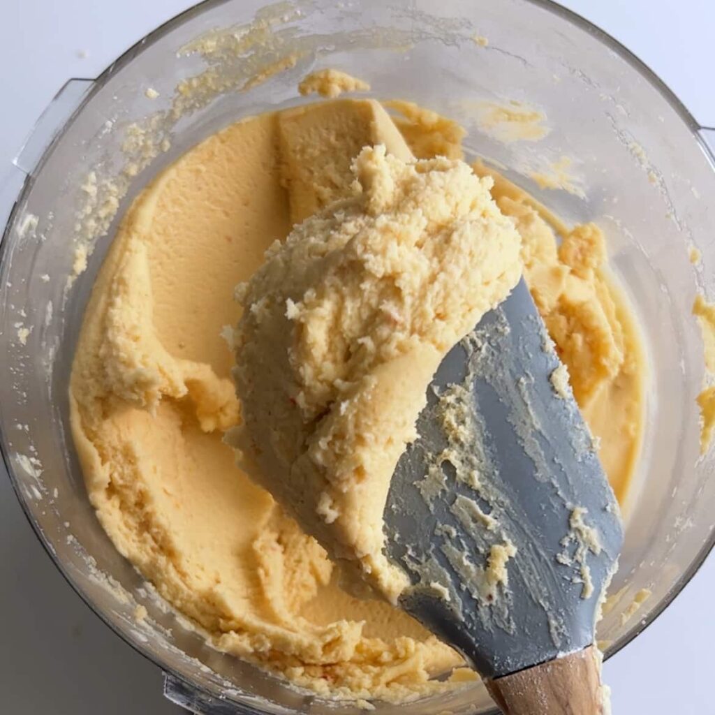 Cheese straw dough being scooped up from a food processor bowl with a gray silicone spatula