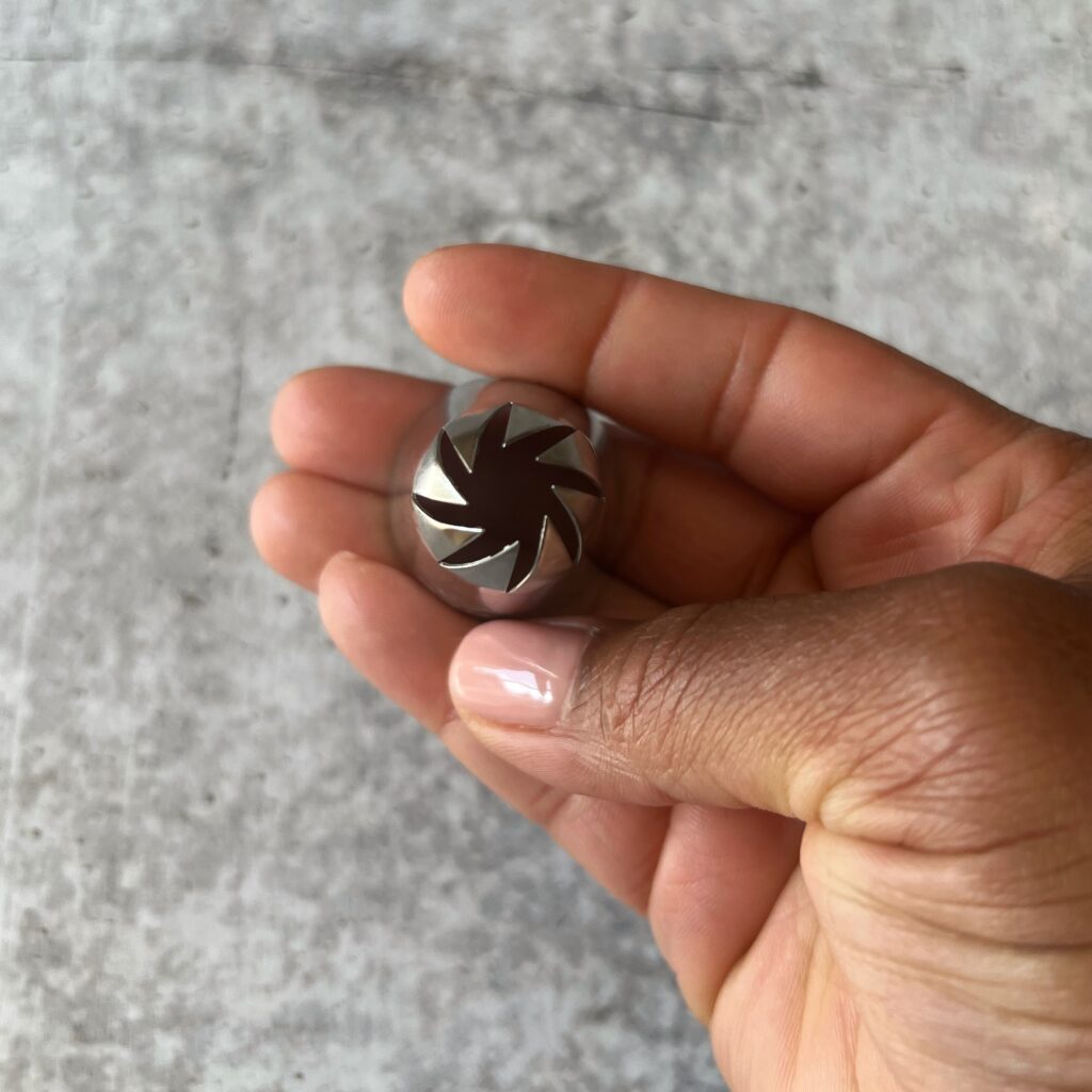 A brown hand holding a Swirl top pastry tip over a concrete background