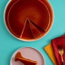 Flan in a pan with a slice of flan on an aqua background. Orange and yellow napkins folded on a green background with a brass and black fork resting on top