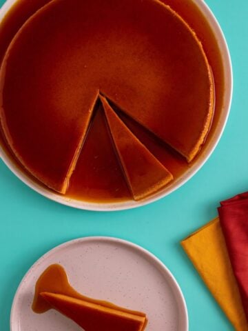 Flan in a pan with a slice of flan on an aqua background. Orange and yellow napkins folded on a green background with a brass and black fork resting on top