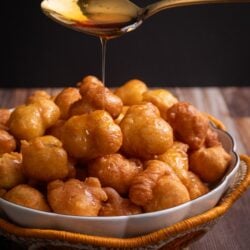A white bowl with a gold rim sitting in a woven basket with an orange scallop edge. The basket is filled with Guyanese Portuguese pancakes. A spoon hovers above the bowl with a long thin pour of syrup being drizzled over the pancakes.