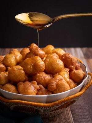 A white bowl with a gold rim sitting in a woven basket with an orange scallop edge. The basket is filled with Guyanese Portuguese pancakes. A spoon hovers above the bowl with a long thin pour of syrup being drizzled over the pancakes.