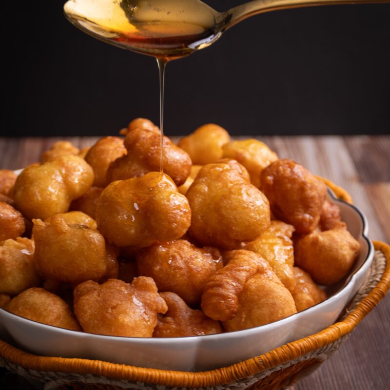 A white bowl with a gold rim sitting in a woven basket with an orange scallop edge. The basket is filled with Guyanese Portuguese pancakes. A spoon hovers above the bowl with a long thin pour of syrup being drizzled over the pancakes.