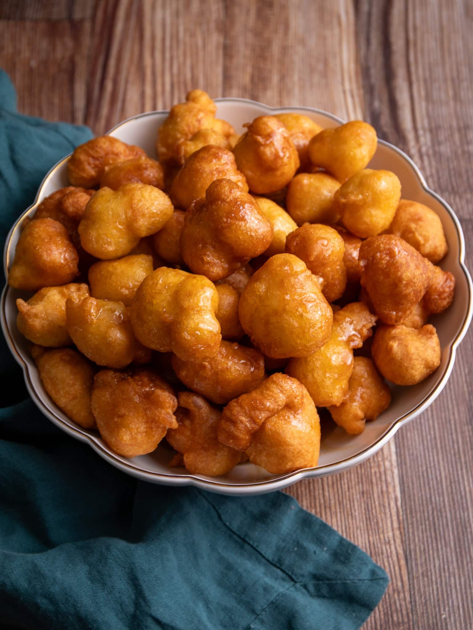 A white bowl with a gold scalloped edge filled with Portuguese Pancakes (malasadas) on a wooden background with a green napkin to the left of the image.
