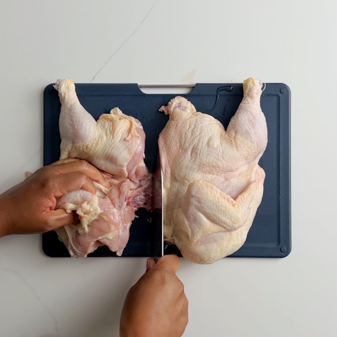 whole chicken cut in half on a blue cutting board. Brown hands holding a knife in between the chicken halves in a slicing motion.