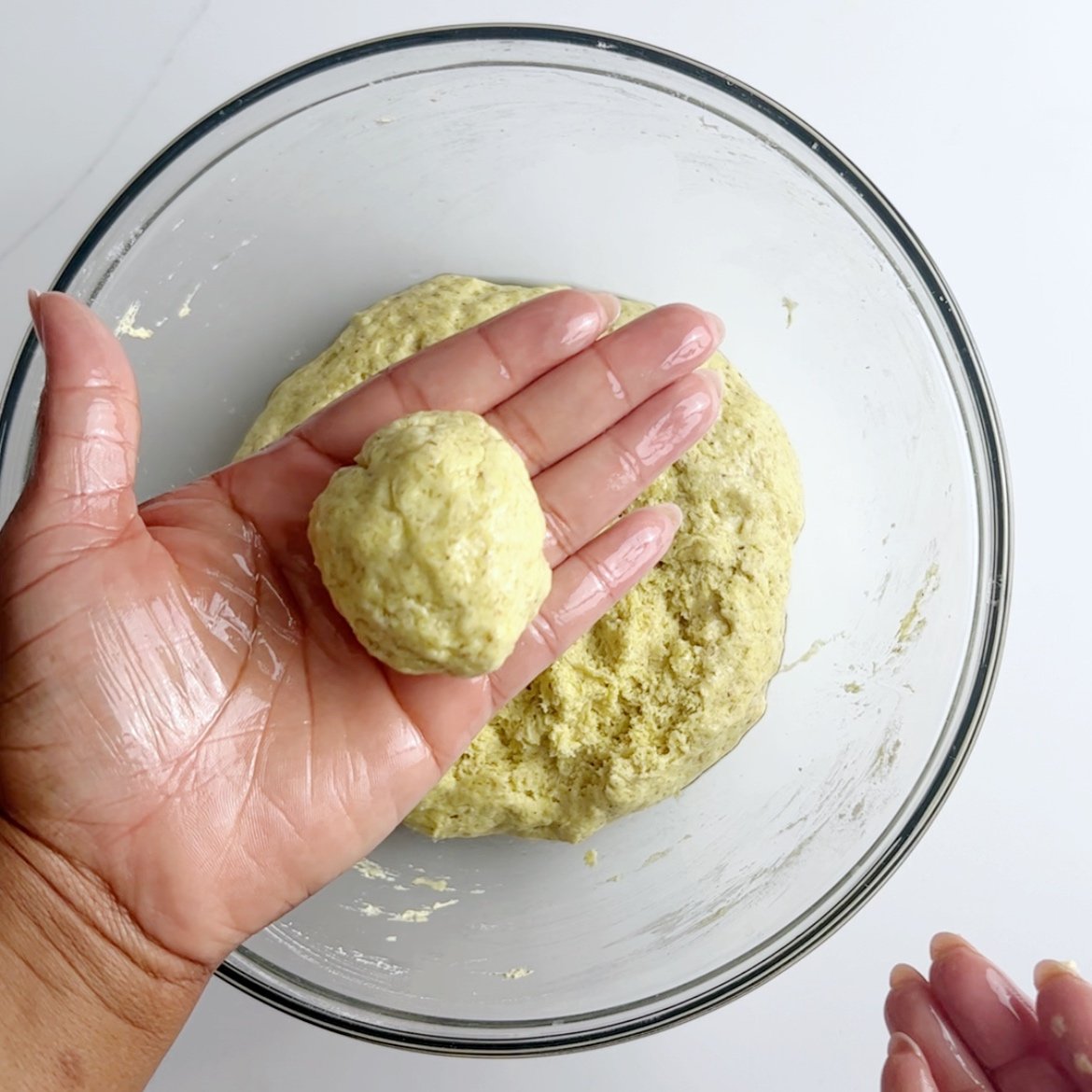 brown hand holding a ball of gluten free bara dough over a glass bowl with more dough