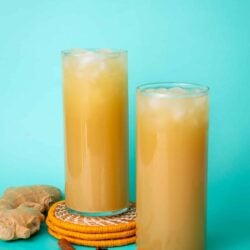 Ginger beer poured into two glasses. One glass is stacked on orange coasters and the other is flat. The background is aqua colored and there are cloves and cinnamon around the glasses.