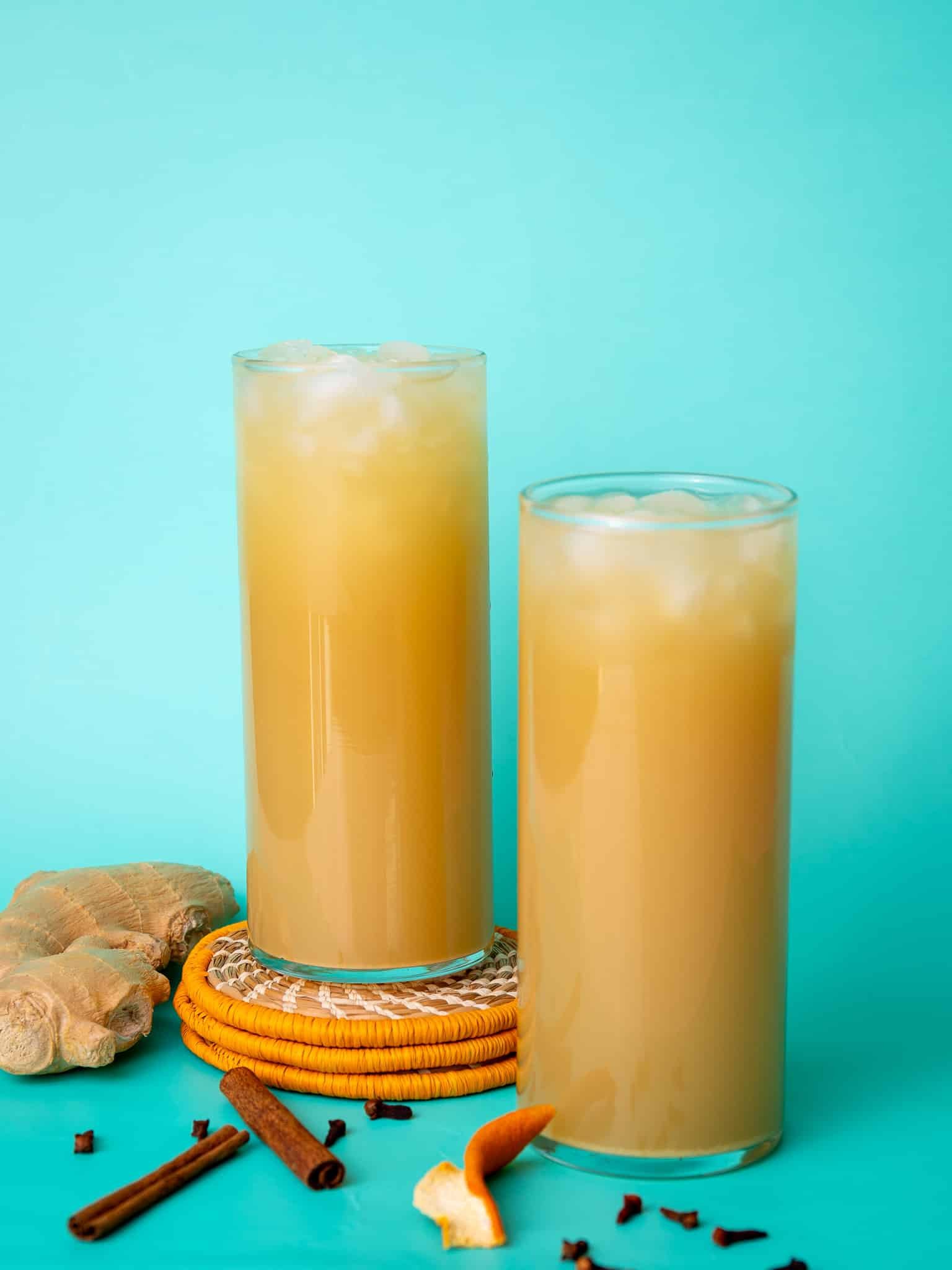 Ginger beer poured into two glasses. One glass is stacked on orange coasters and the other is flat. The background is aqua colored and there are cloves and cinnamon around the glasses.