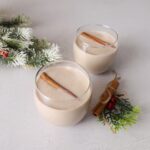 Two glasses filled coquito with cinnamon sticks floating to the top. The glasses are on a white counter with some christmas decorations to the right and left.