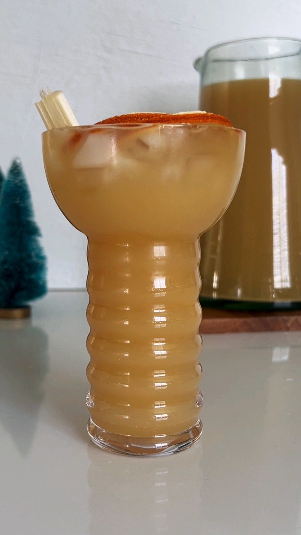 Guyanese ginger beer in a swirly base glass with a slice of orange visible at the top and a mug of ginger beer in the background