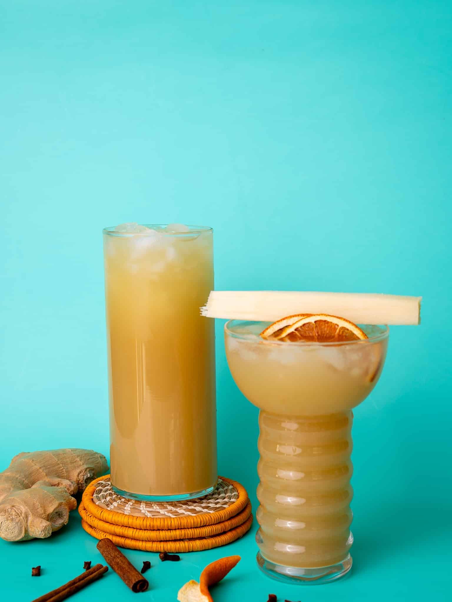 Ginger beer poured into two glasses. One glass is stacked on orange coasters and the other is flat. The background is aqua colored and there are cloves and cinnamon around the glasses.