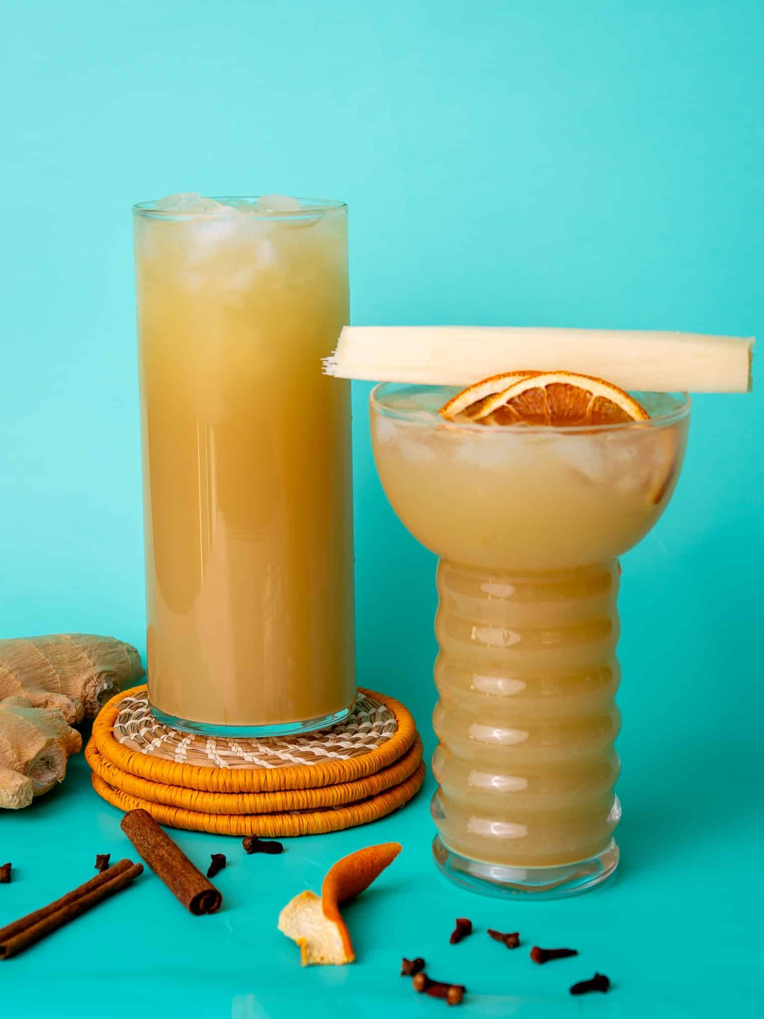 Two glasses of ginger beer on a aqua background. One is topped with a piece of cane sugar and the other is stacked on orange coasters