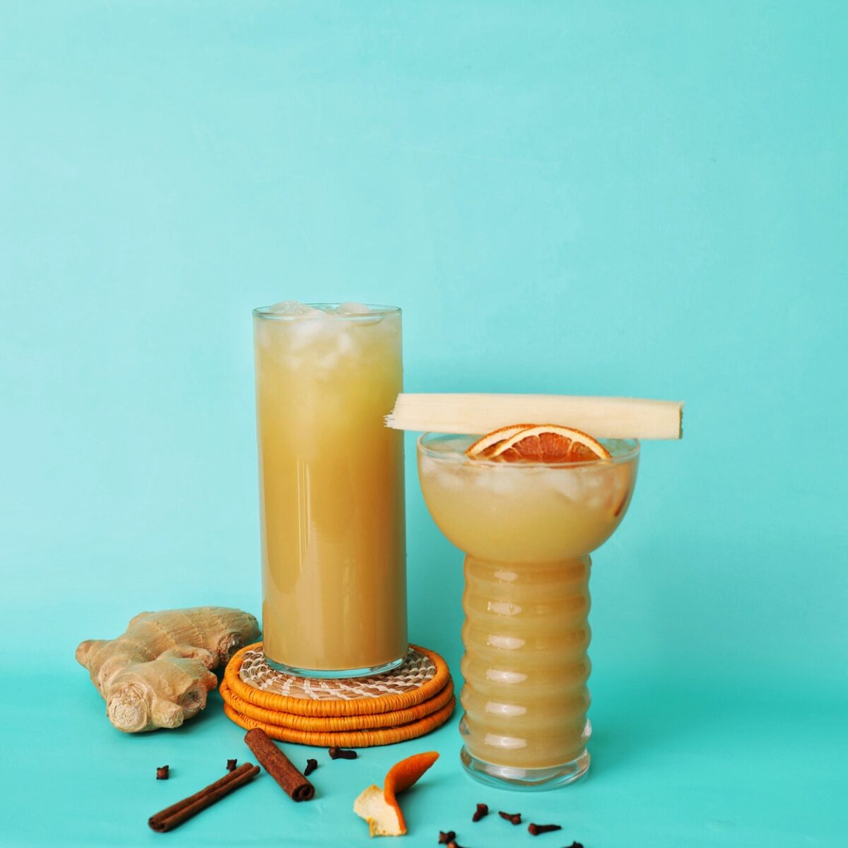 two glasses of ginger beer on a teal black ground. One glass is sitting on a stack of orange coasters. The other glass is topped with a piece of sugar cane, 