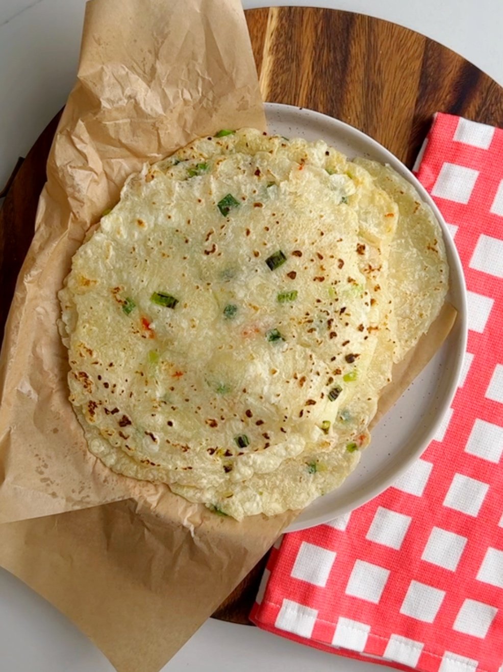 Cassava rotis stacked on a plate lined with parchment paper and a checkered kitchen towel to the side.